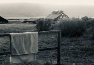 a black and white photo of an old barn above a soaking pool in eastern Oregon. A single towel hangs over a fence post.
